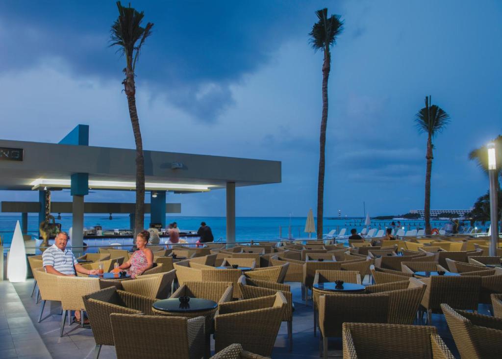 Romantisks vakars pludmales restorānā ar skatu uz jūru, kur pāris sēž pie galda, apkārt ir krēslu rindas un palmas.