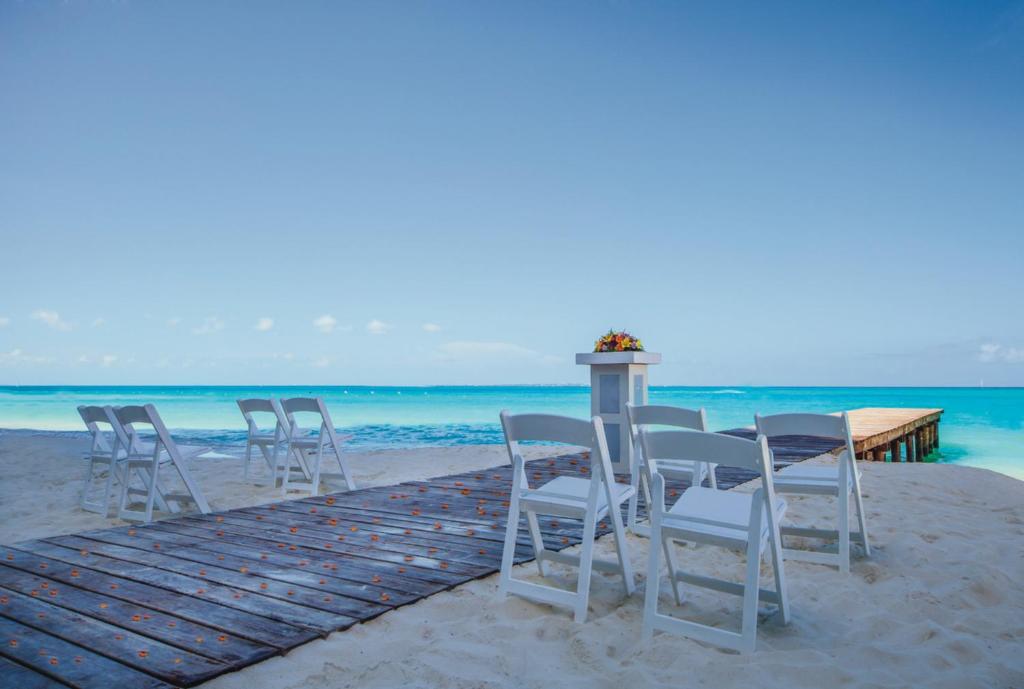 Pludmales kāzu ceremonija ar baltām krēslām un ziedu kompozīciju pie altāra, kas atrodas blakus skaistam zilam ūdenim un koka piestātnei.