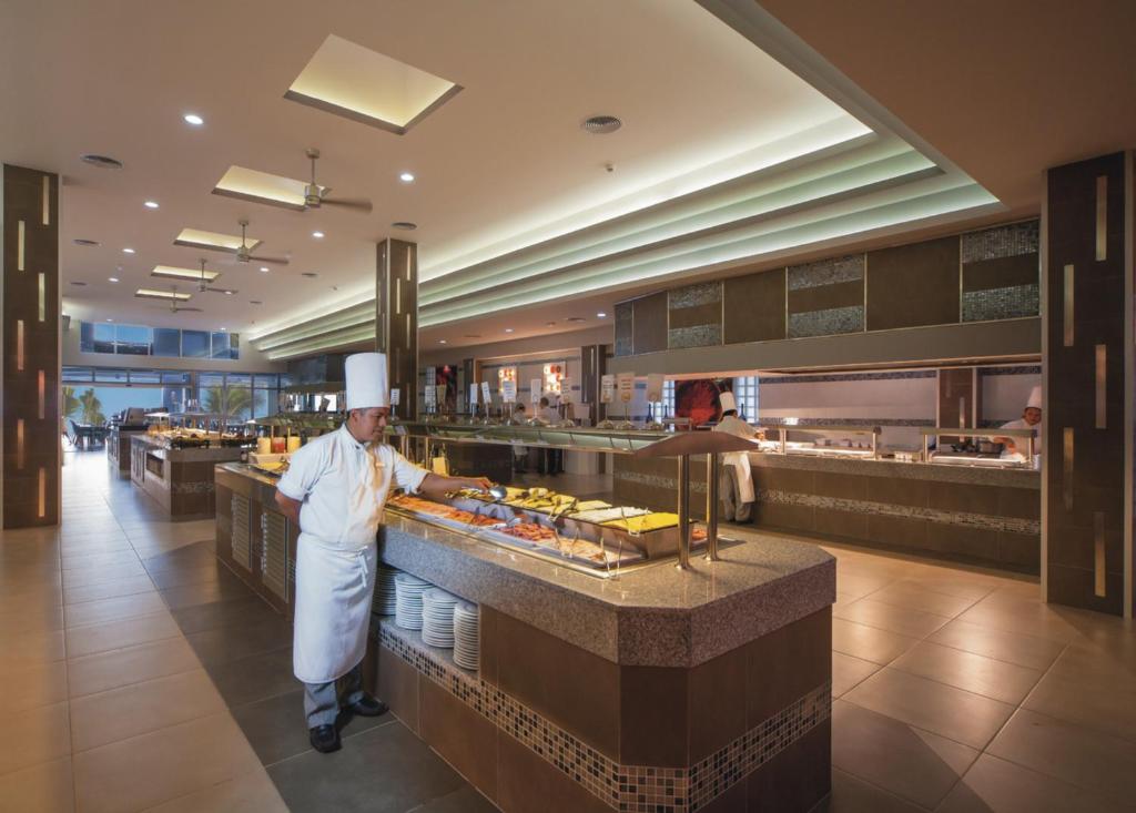 Mūsdienīga restorāna iekārtojums ar šefpavāru, kurš pasniedz ēdienus bufetē. Gaismas un plaša telpa veido patīkamu atmosfēru.