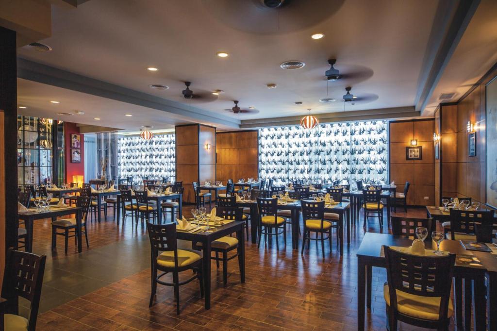 Mūsdienīgs restorāns ar elegantu interjeru, kas iekārtots ar koka mēbelēm un siltu apgaismojumu, radot patīkamu atmosfēru. Galdi ir sakārtoti, gaidot apmeklētājus.