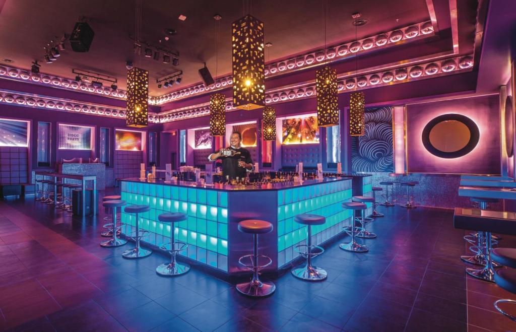 Moderns nakts klubs ar spilgtām gaismām un elegantu bāra letes dizainu, kurā apkalpo bārmenis. Telpa ir dekorēta ar krāsainiem elementiem un mūsdienīgu atmosfēru.