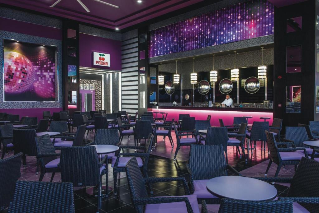 Mūsdienīgs bārs ar purpura un melnā krāsā, stilīgām mēbelēm un izsmalcinātu apgaismojumu, kas rada dzīvīgu atmosfēru. Bāra letē redzams barmenis, kas pasniedz dzērienus.