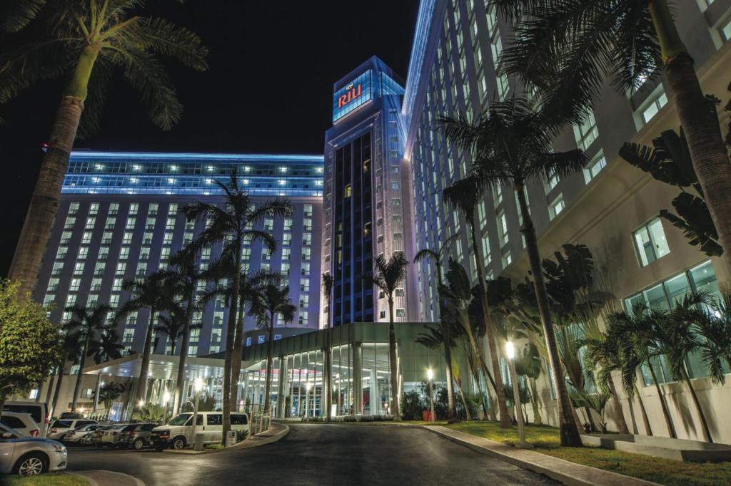 Moderns viesnīcas ēkas skatījums naktī, apgaismots ar krāsu gaismām un ieskauts palmām. Viesnīca ir Riu zīmola.