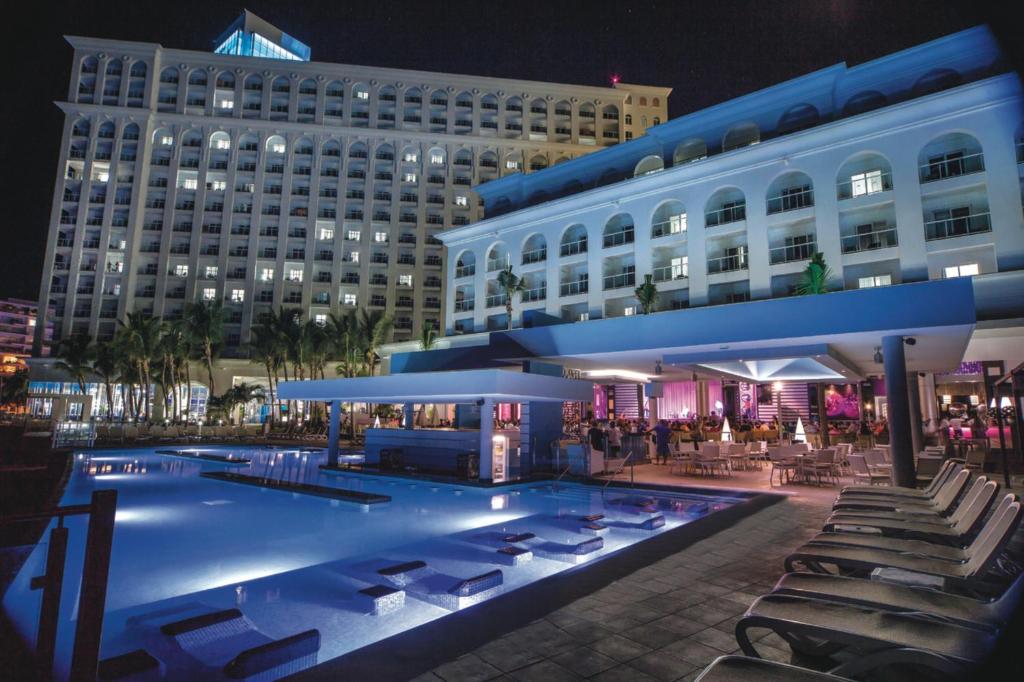 Moderns viesnīcas komplekss ar baseinu un nakts apgaismojumu, kurā redzamas viesu ērtības un apkārtējās palmas. Ideāla vieta relaksācijai un izklaidei.