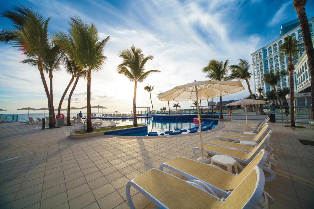 Saulaina pludmales kūrorta ainava ar palmām un baseinu, kas ir apņemts ar sauļošanās krēsliem un lietussargiem. Fons ietver skaistu debesi un viesnīcu.