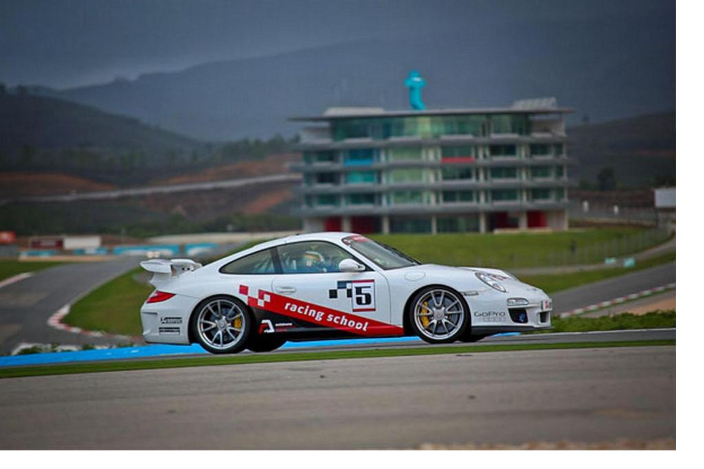 Baltā sacīkšu automašīna ar uzrakstu "racing school" brauc pa trasi, kamēr fons veido kalnu ainavu un modernu ēku.