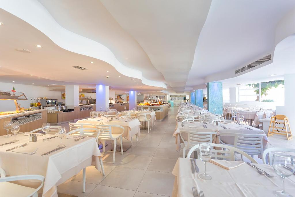 Mūsdienīgs restorāns ar gaišu interjeru, kurā redzamas eleganti klātas galdi un plašas telpas. Apgaismojums rada patīkamu atmosfēru, ideāli piemērotu maltītēm.