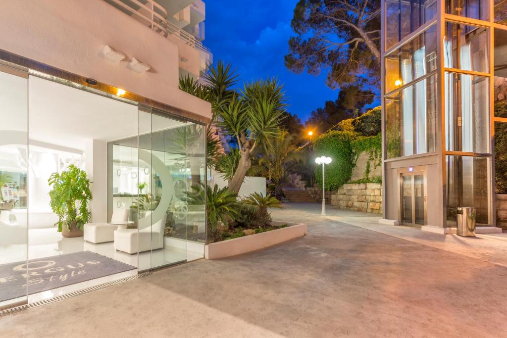 Mūsdienīgs viesnīcas ieeja ar stikla durvīm un augstiem palmām, kas apgaismota nakts gaismā. Blakus redzams stikla lifts un moderna apzaļumota zona.