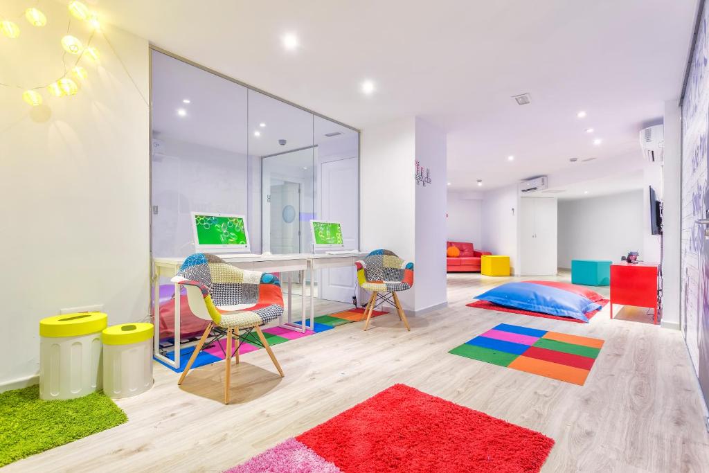 Krāsains un modernais bērnistabas interjers ar datora galdiem, spilgtām krāsām un mīkstiem paklājiem. Telpā ir dažādas sēdvietas un rotaļu zonas, radot draudzīgu un radošu atmosfēru.