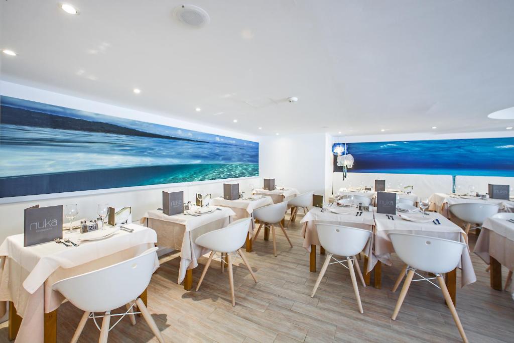 Mūsdienīgs restorāns ar gaišu interjeru un jūras ainavu uz sienas, kur galds ir klāts ar elegantiem galda piederumiem un dekoratīvām aploksnēm.