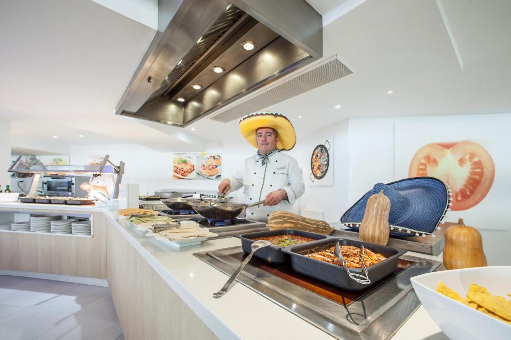 Šefpavārs ar lielu sombrero gatavo ēdienus modernā restorānā, kur redzami dažādi ēdieni un sastāvdaļas. Gaisma ir spilgta un telpa izskatās viesmīlīga un moderna.