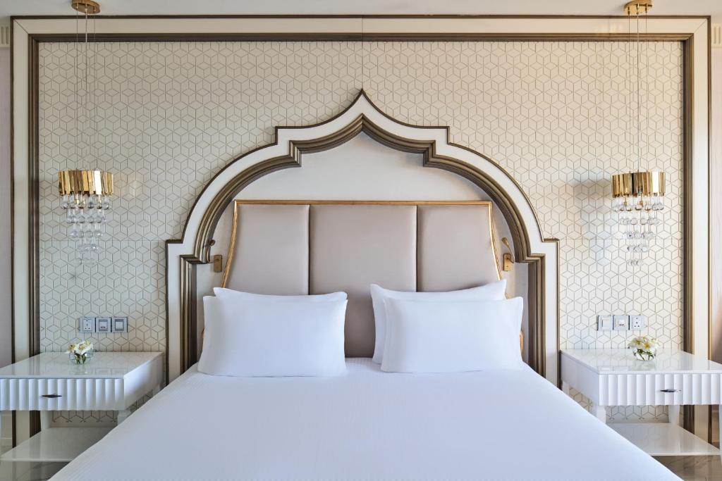 Elegants guļamistabas interjers ar izsmalcinātu gultas galvu, modernām lampām un ģeometrisku tapeti. Mierīga atmosfēra, kas aicina uz atpūtu.