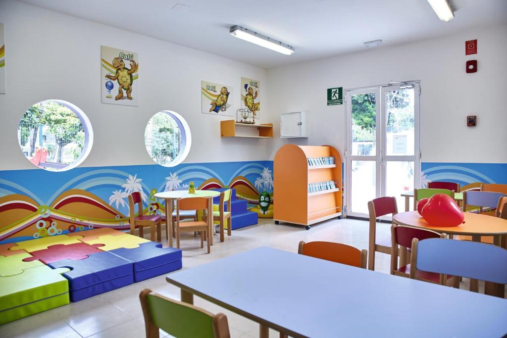 Bērnudārza telpa ar krāsainiem galdiem un krēsliem, spilgtām sienām un rotaļu zonu. Logi ar apaļām formām un bērnu grāmatu plaukts papildina draudzīgo atmosfēru.