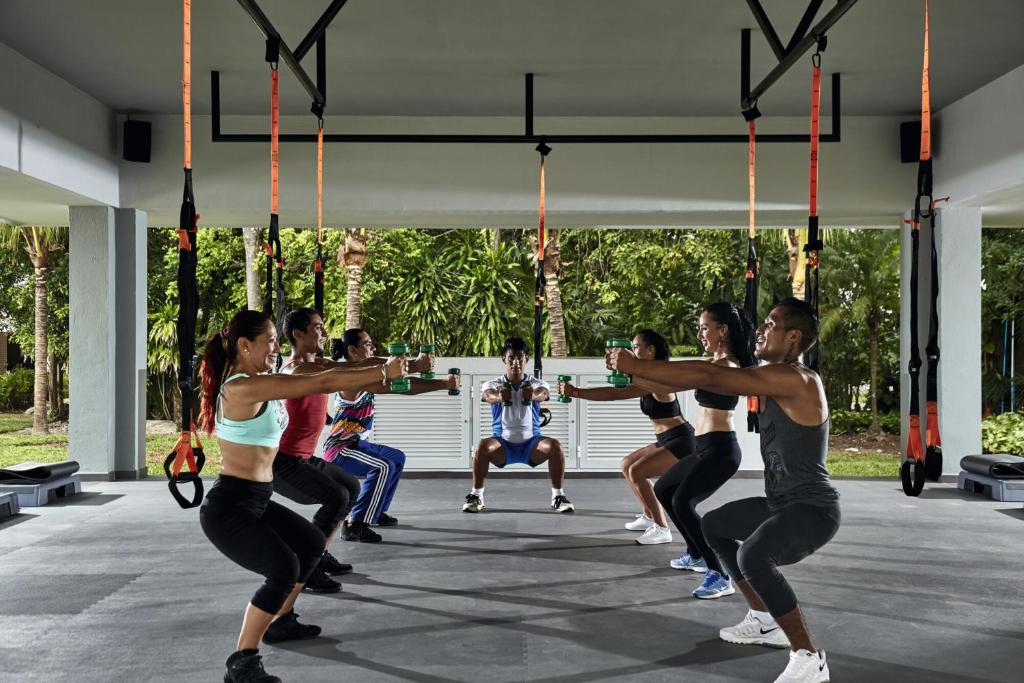 Grupas treniņš sporta zālē, kas ietver dažādas vingrošanas pozas ar hantelēm, apzaļumotā vidē. Sportisti strādā pie savas fiziskās sagatavotības un spēka.