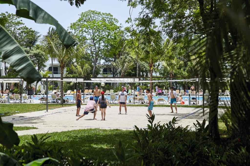 Pludmales volejbola spēle starp grupu cilvēku, fonā redzams baseins un tropiskas augu ainavas.