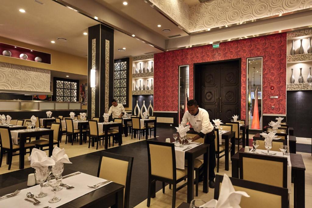 Elegants restorāns ar modernu interjeru, kur viesmīļi sagatavo galdus ar izsmalcinātām rotājumiem. Pievērsta uzmanība detaļām un atmosfērai.