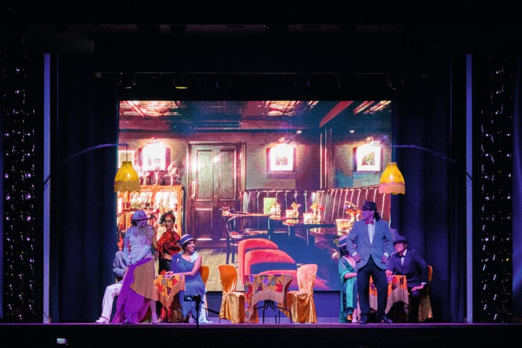 Teātra izrāde ar aktieriem uz skatuves, fona attēlā redzama stilizēta kafejnīca. Aktieri tērpti vintage apģērbā, izsaka dramatiskas emocijas un mijiedarbojas viens ar otru.