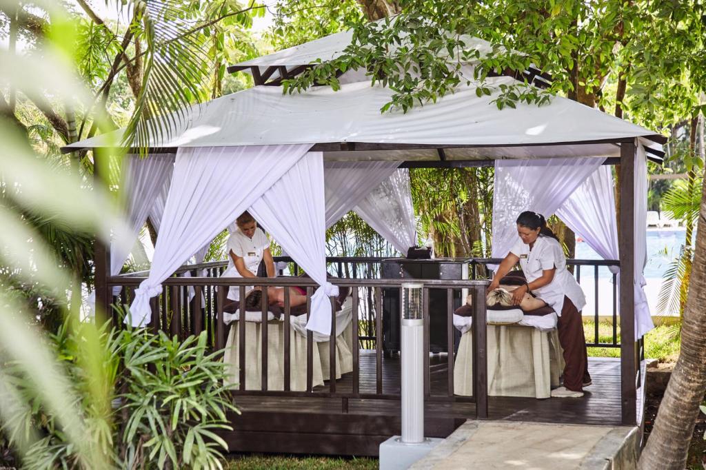 Atpūtas zonas paviljons ar masāžas galdiem, kur divi masieri sniedz pakalpojumus dārzā ar zaļumiem un ērtu atmosfēru.