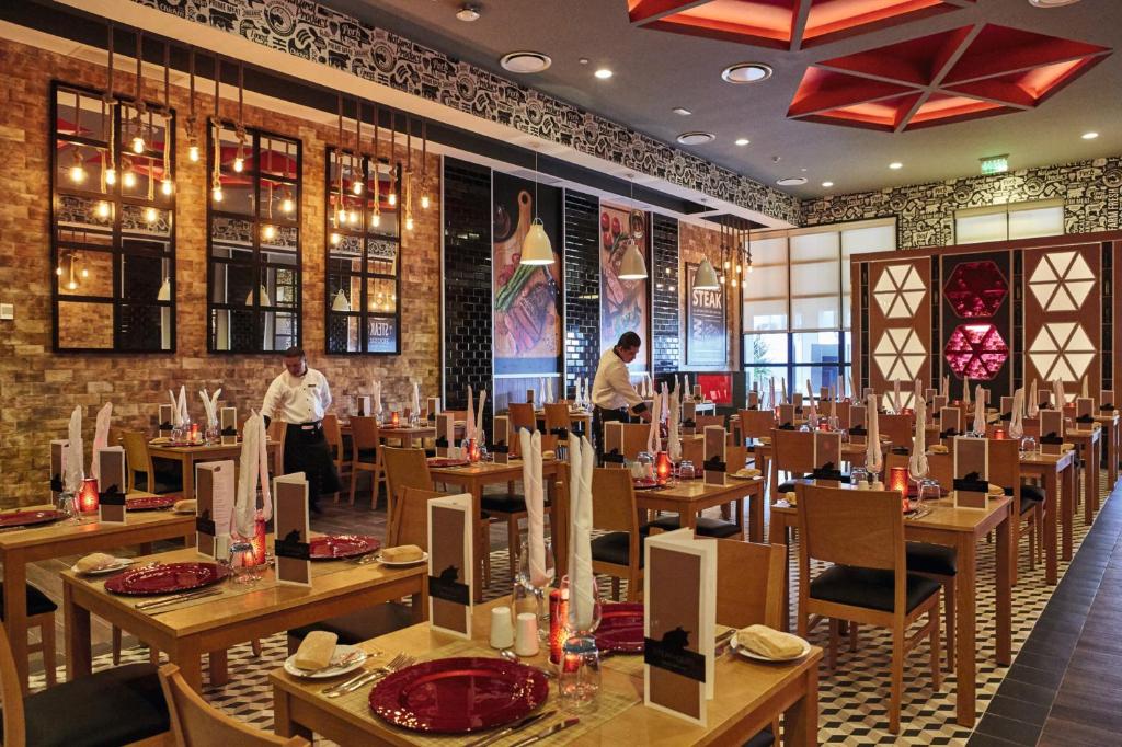 Mūsdienīgs restorāna interjers ar elegantiem galdiem, siltu apgaismojumu un viesmīli, kas gatavo vietu apmeklētājiem. Galdi ir klāti ar sarkaniem šķīvjiem un svinīgiem salvetēm.