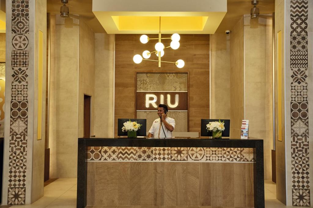 Reģistratūra modernā viesnīcā ar stilīgu dizainu un viesmīlīgu darbinieku, kas runā pa tālruni. Interjerā ir grezni dekori un ziedu kompozīcija.