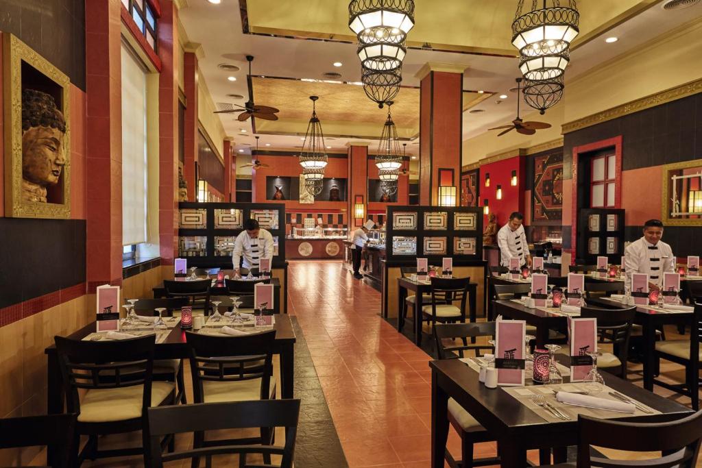 Elegants restorāns ar tradicionālu dekoru un apkalpojošo personālu, kas sagatavo galdiņus. Interjers ir veidots ar siltām krāsām un izsmalcinātu apgaismojumu, radot patīkamu atmosfēru.