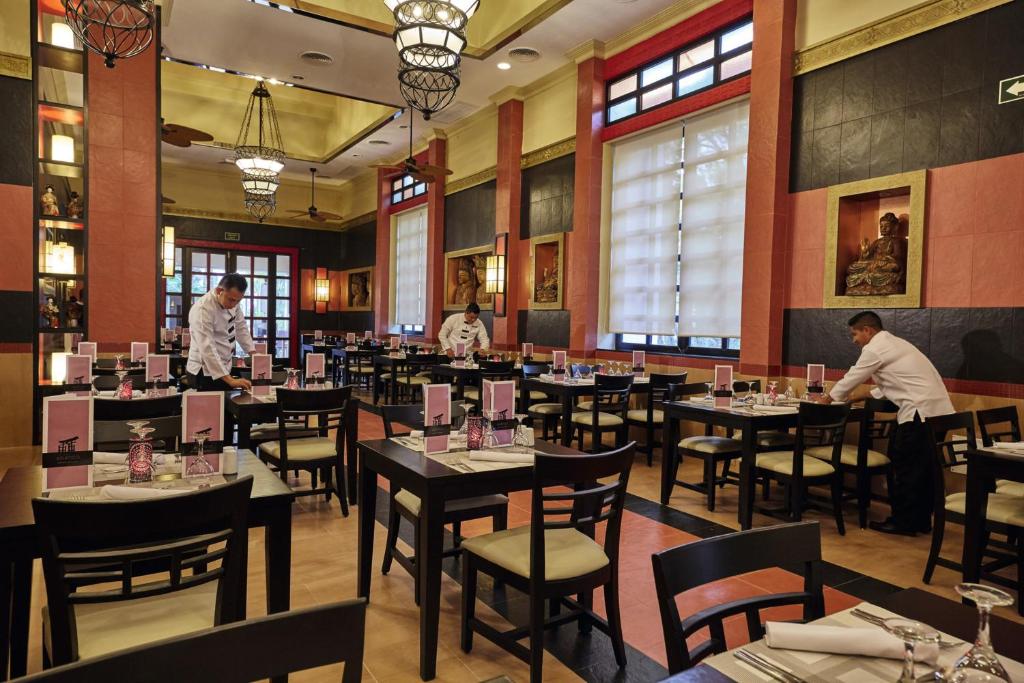 Restorāna interjers ar apkalpojošo personālu, kas sagatavo galdiņus pirms viesu ierašanās. Telpā ir eleganta apgaismojuma un dekoratīvu elementu kombinācija, radot patīkamu atmosfēru.