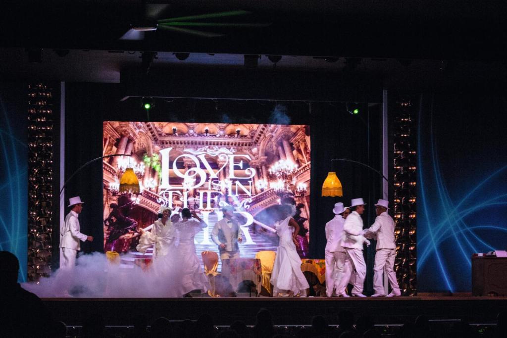 Skatuves izrāde ar dejotājiem baltos tērpos, fonā izceltā vide ar vārdiem 