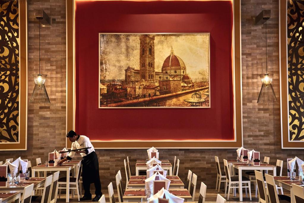 Elegants restorāna interjers ar siltām krāsām, kur apkalpotājs gatavo galdus. Sienā ir gleznains attēls ar Florences arhitektūru, radot patīkamu atmosfēru.