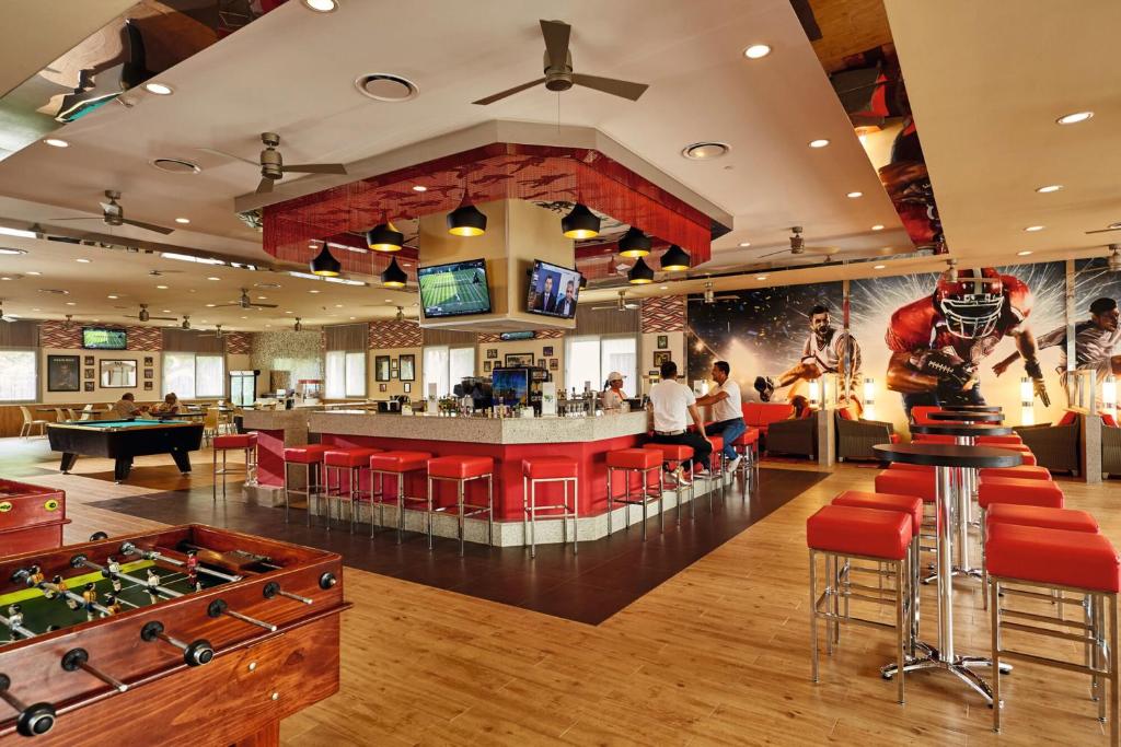 Moderns sporta bārs ar spēļu galdiem, sarkaniem krēsliem un lieliem ekrāniem, kas rāda sporta notikumus. Telpa ir gaiša un dzīvīga, ar sporta tēmu sienām un ērtām sēdvietām.