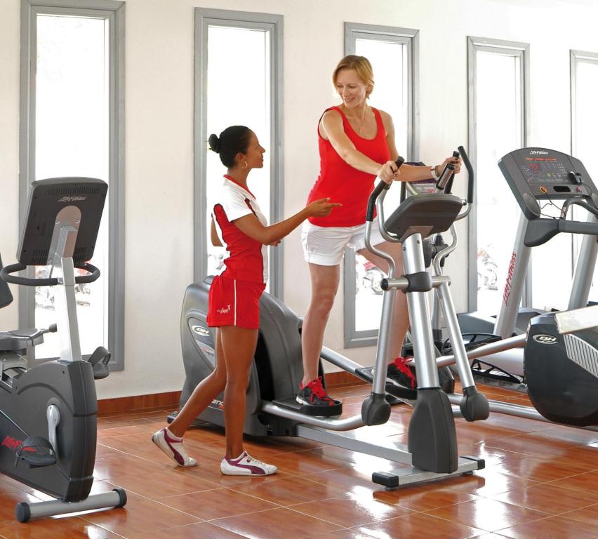 Sieviete sporta zālē saņem apmācību no treneres, izmantojot eliptisko trenažieri, kamēr apkārt ir dažādi sporta aprīkojumi.