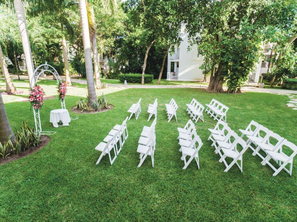 Labi iekārtota kāzu ceremonijas vieta ar baltiem krēsliem un ziedu dekorēto arku, zaļā zālē, apstādījumos. Ideāla vide svinībām.