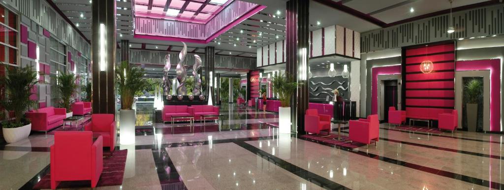 Mūsdienīgs viesnīcas vestibils ar rozā un melnā krāsā veidotu interjeru, kurā ir eleganti dīvāni un dekoratīvi augi. Gaisma piešķir telpai dinamisku un stilīgu atmosfēru.