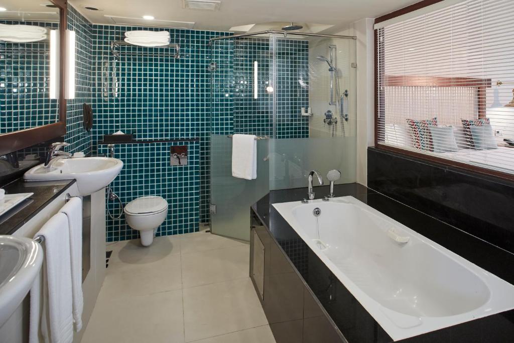 Mūsdienīgs vannas istabas interjers ar tumši zaļiem mozaīkas flīzēm, vannu, dušas kabīni un uzglabāšanas vietām. Elegants dizains un funkcionālas iekārtas.