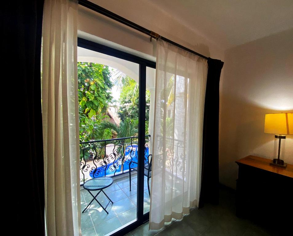 Balkona skats ar zaļumiem un baseinu, caur caurspīdīgām aizkaru drānām. Telpa ar lampu un koka mēbeli, radot mājīgu atmosfēru.