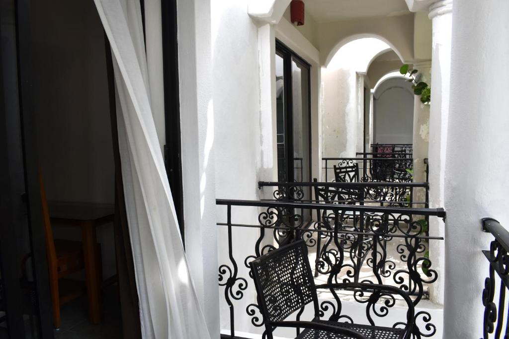 Balconu ar dzelzs margām un krēsliem, kas vērsti uz gaišu, atvērtu telpu. Gaisma ieplūst caur logiem, radot patīkamu atmosfēru.