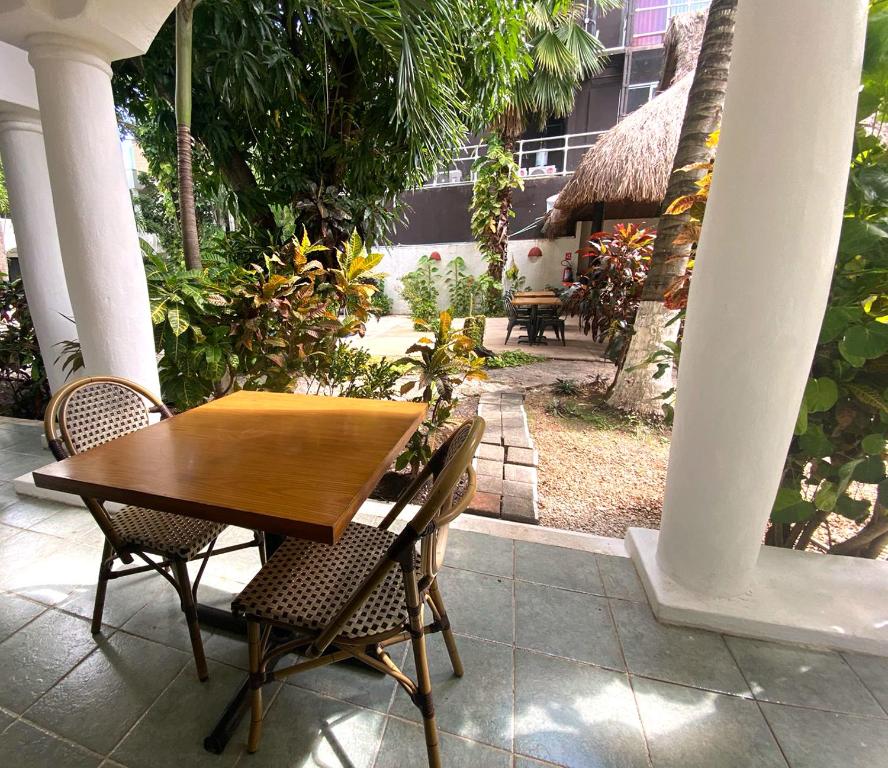 Āra terase ar koka galdu un krēsliem, ieskauta zaļumiem un augiem. Fons ietver brīvdabas ēdamzāli un dārzu, radot mierīgu atmosfēru.