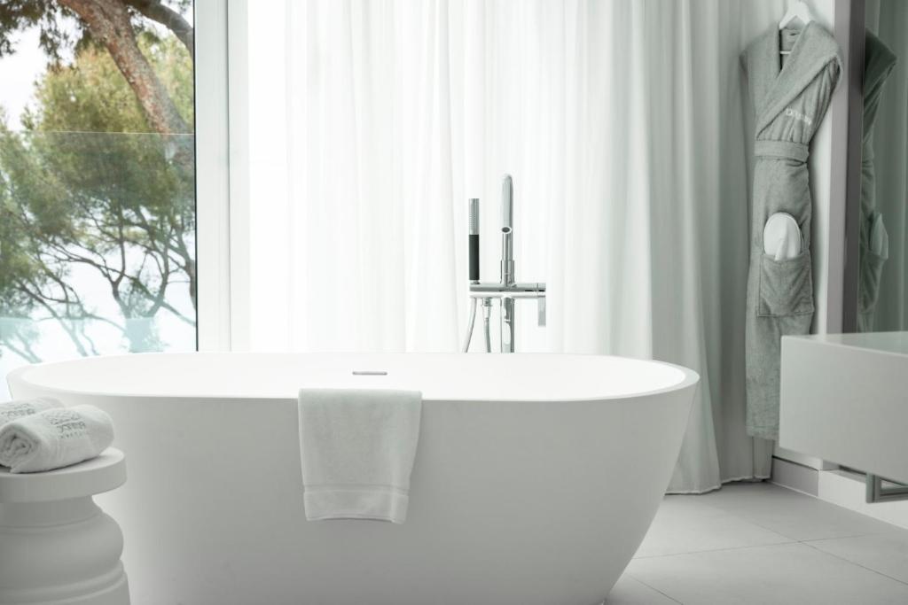 Moderns vannas istabas interjers ar elegantu brīvi stāvošu vannu, baltām dvielēm un mīkstu halātu, kas piekārts uz āķa. Lielie logi nodrošina dabīgu apgaismojumu un skatu uz dabu.