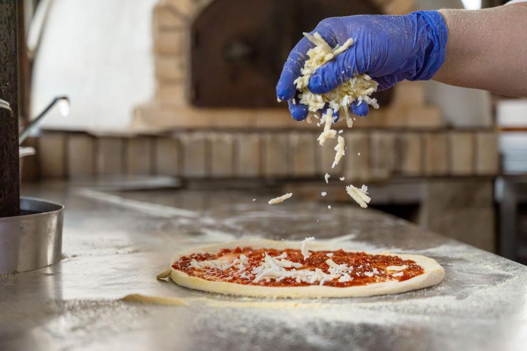 Siera izklāšana uz picas ar tomātu mērci, kamēr tiek izmantotas zilas cimdi. Fons atspoguļo picas cepšanas vidi.