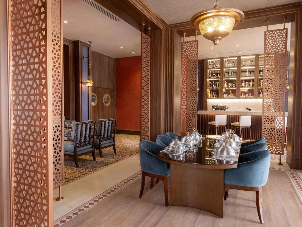 Elegantā restorāna interjers ar sudraba traukiem uz galda, izsmalcinātiem zilu krēslu komplektiem un piesātinātu koka apdari. Redzams arī bārs ar plašu dzērienu izvēli fonā.