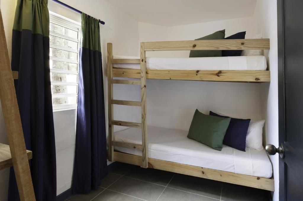 Mūsdienīga guļamistaba ar divstāvu gultu, kas veidota no koka, un saskaņotām spilveniem. Logā ir vertikāli žalūzijas un tumši zili aizkari, radot mājīgu atmosfēru.