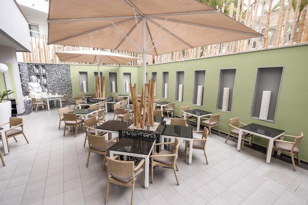 Moderns restorāns ar ērtām ratiņkrēslām un melniem galdiem, kas izvietoti zem liela saules jumta. Fons veidots ar zaļām sienām un dekoratīviem elementiem, radot relaksējošu atmosfēru.