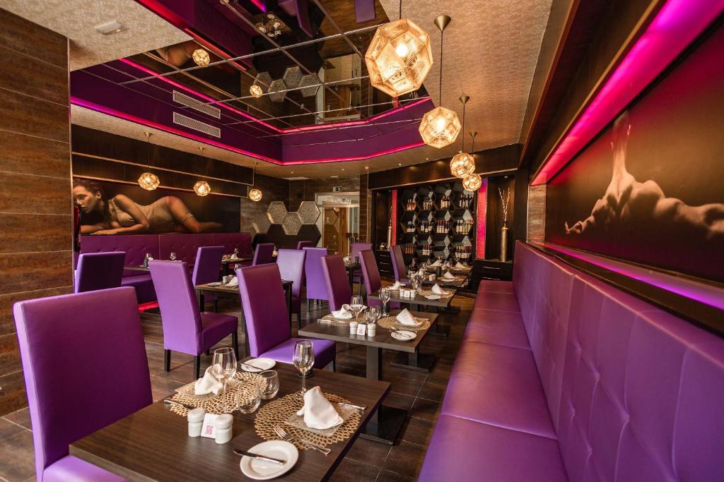 Mūsdienīga restorāna interjers ar purpura krāsas mēbelēm, stilīgām lampām un elegantām galda uzstādījumiem. Gaisotne ir modernā un pievilcīgā dizainā, radot patīkamu atmosfēru.