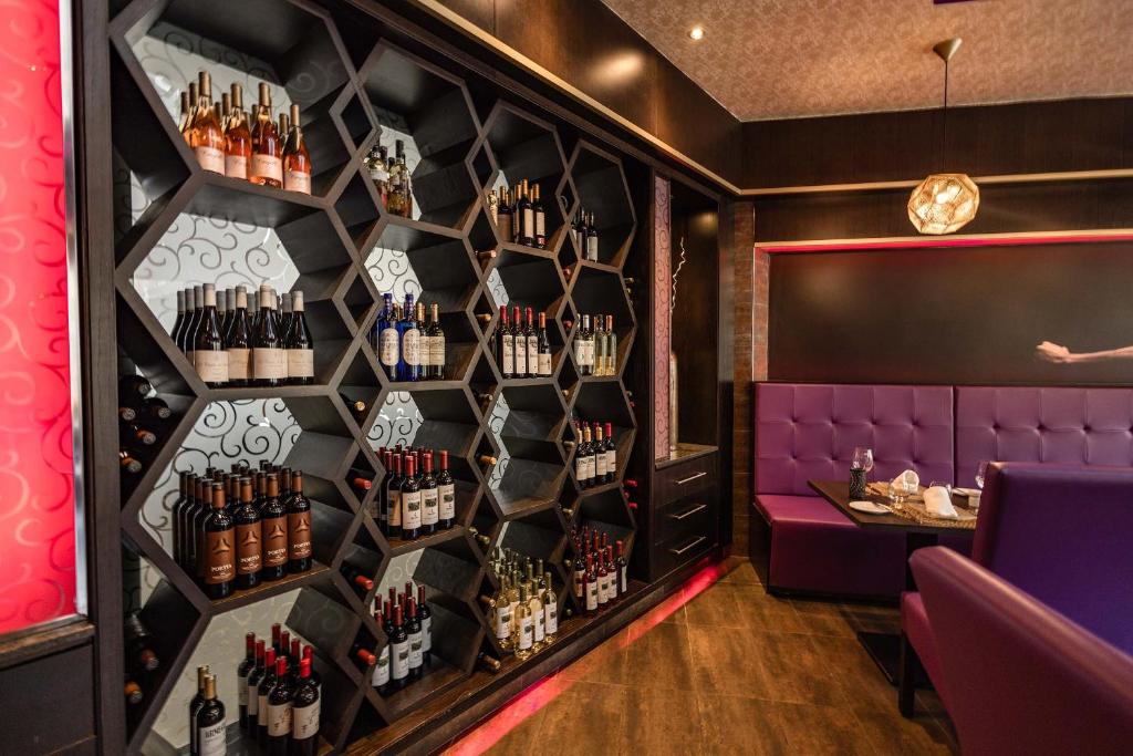 Mūsdienīgs vīna plaukts ar sešstūra formu, kas satur dažādas vīna pudeles, un elegantas sēdvietas restorānā ar purpursarkanām krēsliem.