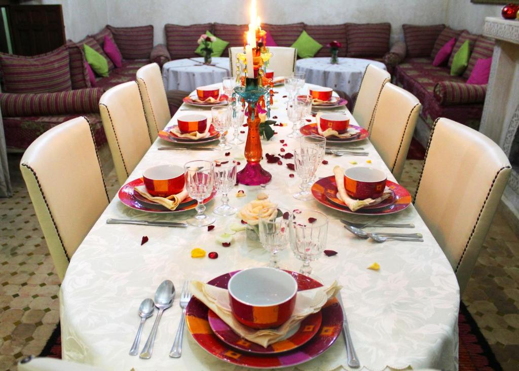 Eleganti izklāta ēdamzāle ar krāsainiem galda piederumiem, svečturi un ziedlapiņām uz galda, radot svinīgu atmosfēru.