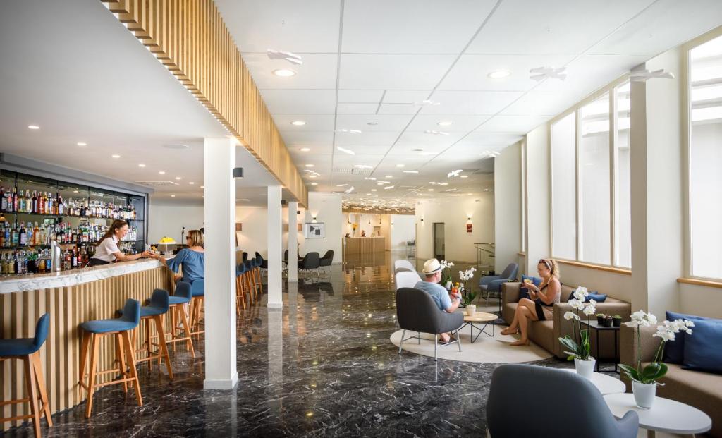 Mūsdienīgs bārs ar elegantu interjeru, kurā cilvēki atpūšas un izbauda dzērienus. Gaismas un plašums rada viesmīlīgu atmosfēru.