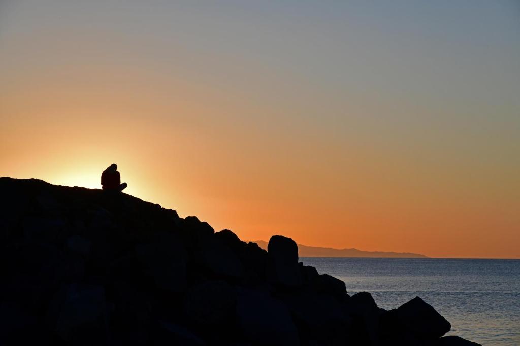 Saulrieta ainava ar cilvēku, kas sēž uz akmeņu krāvuma jūras malā. Siluets pret debesīm ar siltām oranžām un dzeltenām krāsām.