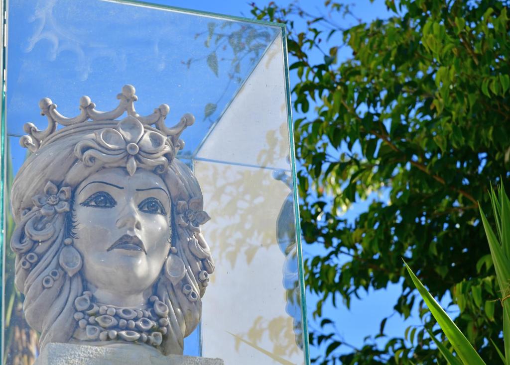 Mākslas skulptūra, kurā attēlota sievietes seja ar kroni un rotājumiem, ir izvietota stikla vitrīnā, fonā redzami zaļi koki un skaidra zila debess.