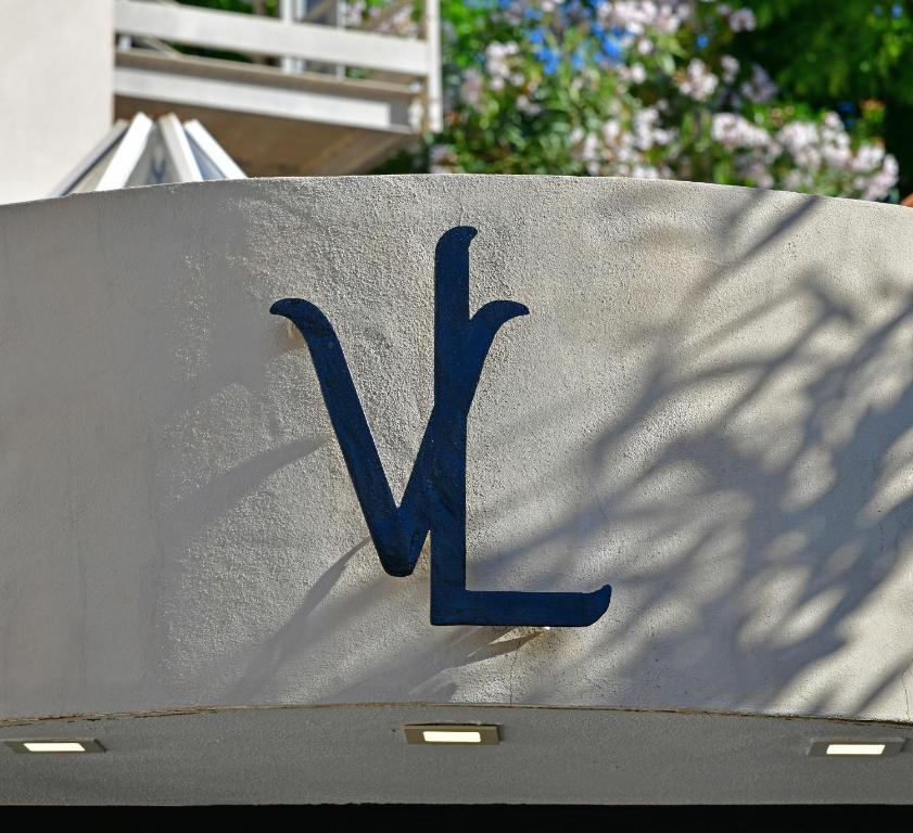 Melna logo ar burtiem "VL" uz gaišas sienas, fons ar ziediem un zaļumiem.