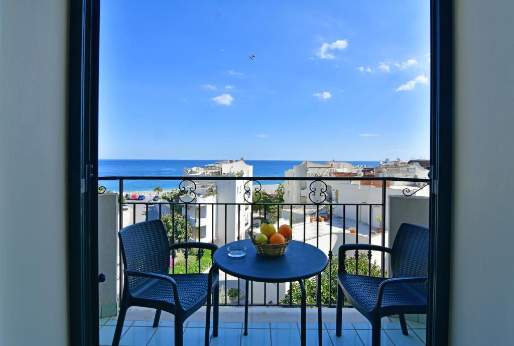 Skats no balkona uz jūru, kurā redzams galds ar augļu grozu un divas krēsli. Fons ir debesis ar dažiem mākoņiem un piekrastes ēkas.