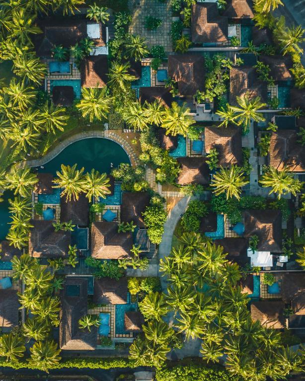 Eksotiska kūrorta ainava ar palmām un peldbaseiniem, redzēta no putna lidojuma. Vairāki tradicionāli veidoti nami, iezaigojuši ainavu ar zaļumu un ūdeni.